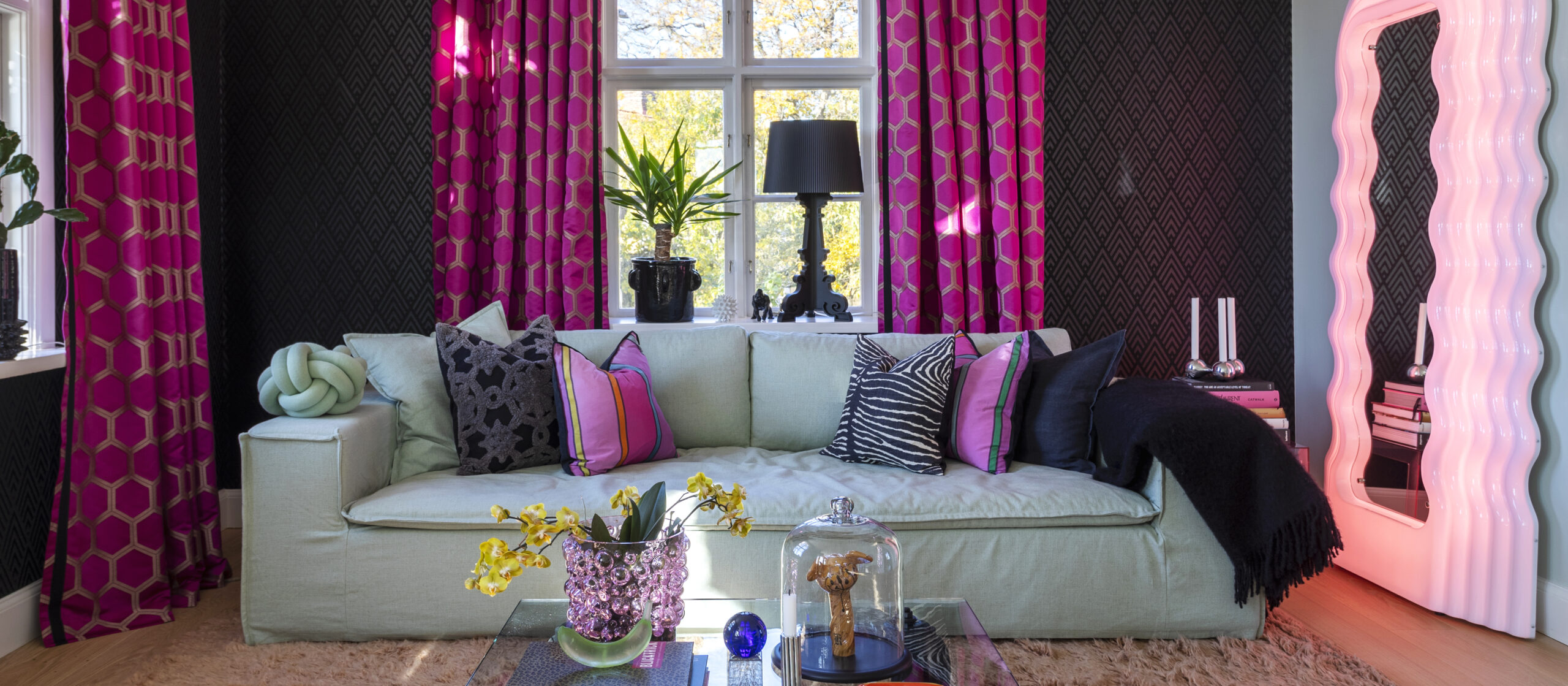 A room with a light green couch, pink wavy mirror, and printed curtains. Colorful pillows and interior pieces complete the decor.